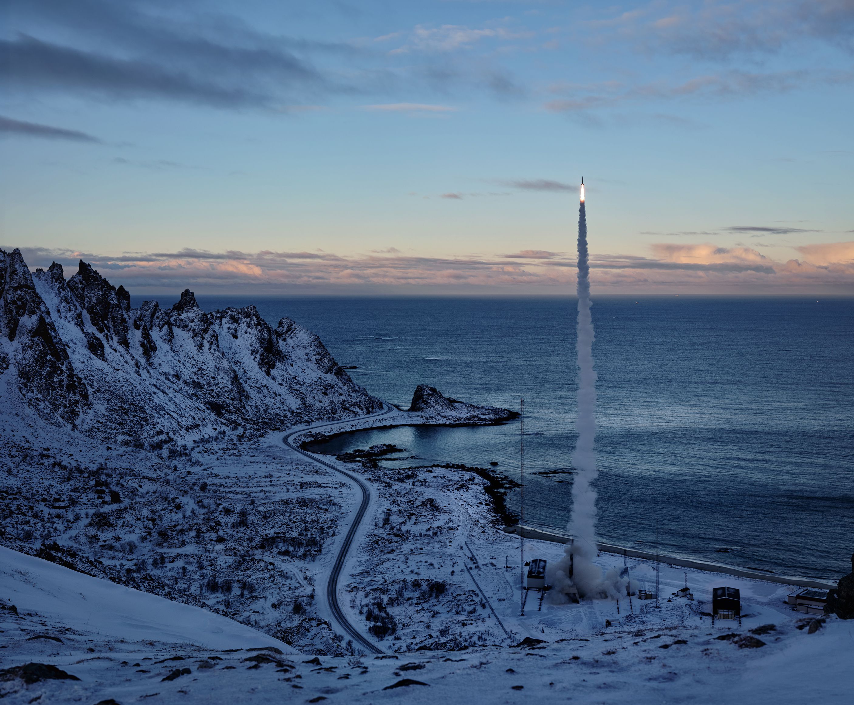 A rocket launches from a coastal base, rising over a snowy landscape and ocean.